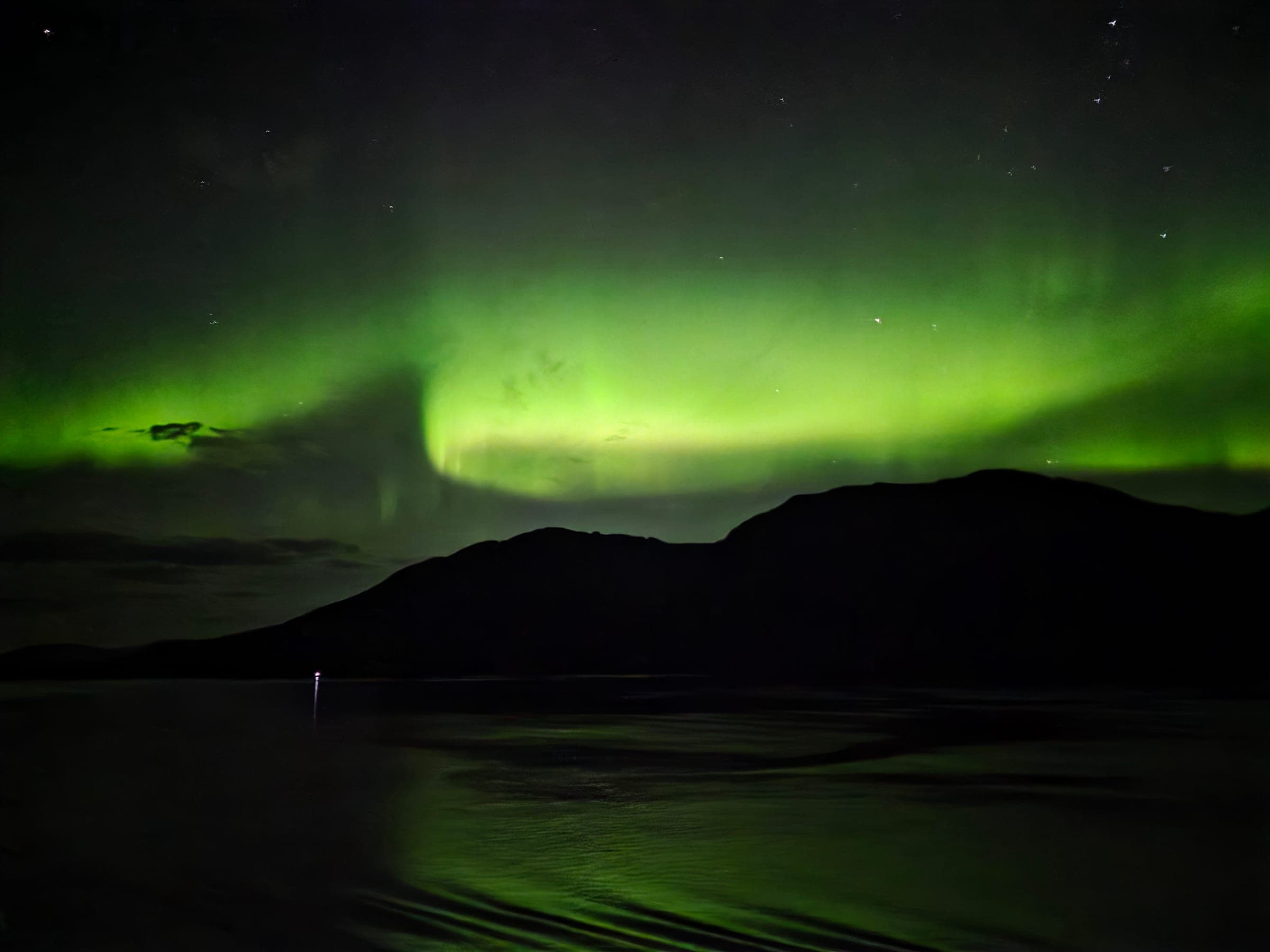 Bright green aurora borealis over a silhouetted mountain ridge, reflected in the water below